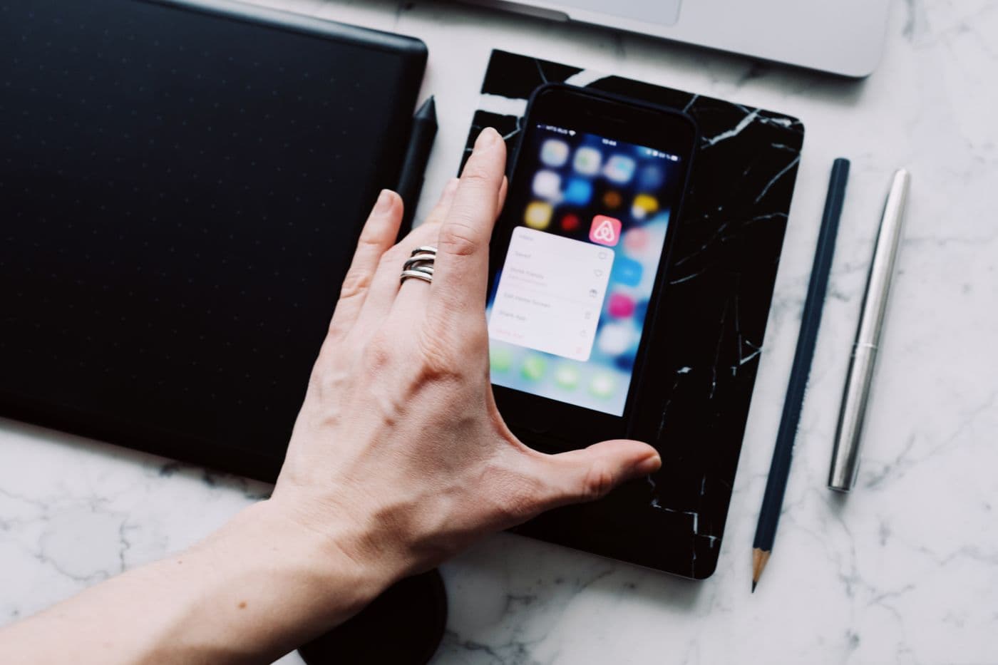 Hand using a smartphone beside a tablet, notebook, and pencils on a marble desk.